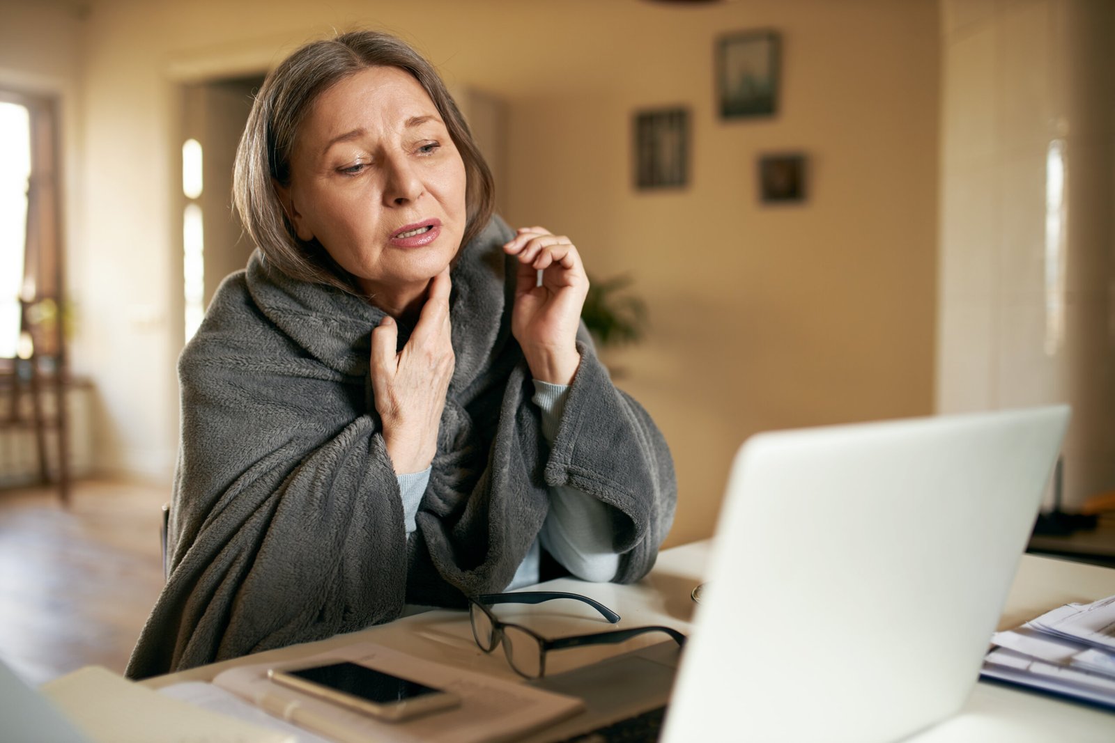 Upset elderly woman talking with doctor via video conference application sitting in front of open laptop, suffering from respiratory disease, complaining of sore throat, having painful look